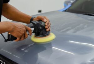 Technician polishing a gray car hood with a buffer.
